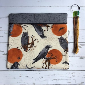 May include: A rectangular fabric zip pouch featuring a cream background with a pattern of black crows, orange full moons, spiderwebs, and spiders. The top section is grey with cursive text reading Haunted, Potions, and Trick. Includes a gold tassel.