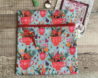 May include: A square, zippered pouch with a teal background and a repeating pattern of red teacups filled with flowers and the word "JOY". The pouch has a red zipper and a red pull. Two small Christmas-themed embellishments are at the top.