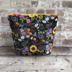 May include: A black floral cosmetic bag with a yellow zipper and pink pull. The bag features a colorful pattern of flowers, butterflies, and inspirational text, including "Don't Worry, Be Happy."