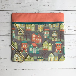 May include: A rectangular fabric pouch featuring a grey background with a repeating pattern of colourful houses, trees, and shops. The top section is solid coral pink, separated by a teal zip with a yellow cord pull. Metal scissors are attached.