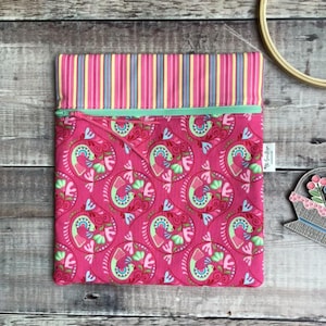 May include: A pink zippered pouch with a colorful floral pattern and a striped top. The pouch has a mint green zipper and a small tag on the side. The design features a repeating pattern of stylized birds and flowers.