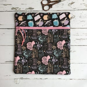 May include: Black book sleeve with a pink zip and pull, patterned with books and reading-themed phrases. The text includes "Shhh!" and "Keep Calm Read On." A pair of scissors is visible above the sleeve.