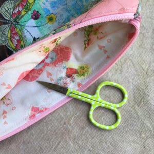 Puede incluir: Una bolsa de tela rosa con una abertura de cremallera y un estampado floral. Un par de tijeras verdes con lunares blancos est&aacute; sobre una superficie marr&oacute;n junto a la bolsa.