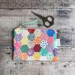 May include: A patchwork hexagon-patterned zipped pouch with a pink zip pull. The pouch features various floral and patterned fabrics. A pair of ornate, vintage scissors rests on the wooden surface beside the pouch.
