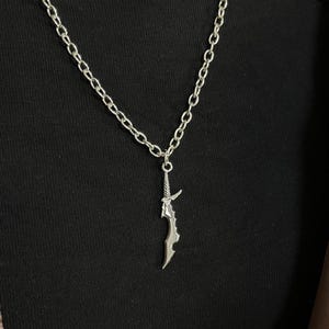 May include: A silver-colored necklace featuring a pendant shaped like a stylized sword. The sword pendant has intricate details and hangs from a chain with oval links. The necklace is displayed against a black background.