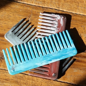 May include: A collection of colorful combs in shades of blue, gray, pink, and brown. The combs are made of a marbled material and have wide teeth. They are arranged on a wooden surface.