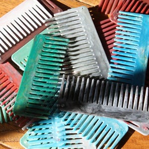 May include: A collection of colorful combs in shades of red, green, blue, gray, and white. The combs are made of plastic and have wide teeth. The combs are scattered on a wooden surface.