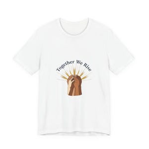 May include: White t-shirt featuring the text "Together We Rise" in a curved font above a graphic of hands of various skin tones. The hands are clasped together, with a sunburst design behind them.