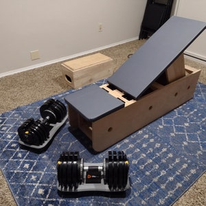 May include: A wooden incline bench with a gray padded top, two adjustable positions, and a wooden box. Two adjustable dumbbells are on a blue and white patterned rug.