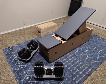 May include: A wooden incline bench with a gray padded top, two adjustable positions, and a wooden box. Two adjustable dumbbells are on a blue and white patterned rug.