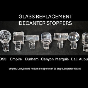 May include: A row of eight glass decanter stoppers in various styles, including a diamond-shaped stopper, a square stopper, and a round stopper. The stoppers are labeled with their names: DS3, Empire, Durham, Canyon, Marquis, Ball, and Auburn. The text "GLASS REPLACEMENT DECANTER STOPPERS" is at the top of the image. The text "Empire, Canyon and Auburn Stoppers can be engraved/personalized" is at the bottom of the image.