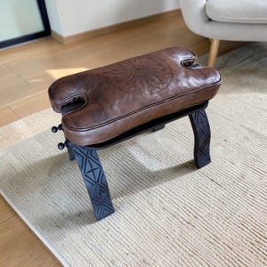 May include: A brown leather bench with a decorative floral pattern on the seat. The bench has a black metal frame with geometric designs on the legs. The bench is approximately 24 inches long, 12 inches wide, and 18 inches high.