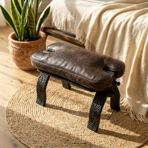 Dark brown leather camel stool cushion on a vintage wooden stool in a cozy home interior, perfect handmade seat replacement for camel saddle stools.
