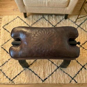 May include: A brown leather ottoman with a carved floral design on top. The ottoman has a rectangular shape with curved sides and black accents. It sits on four dark green legs. The ottoman is placed on a cream-colored rug with a black diamond pattern.