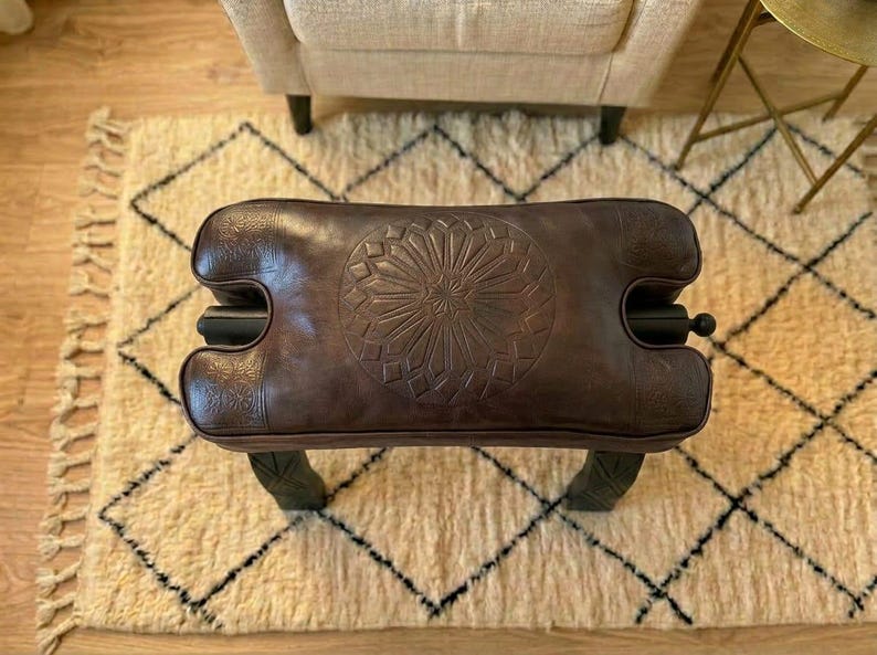 May include: A brown leather ottoman with a carved floral design on top. The ottoman has a rectangular shape with curved sides and black accents. It sits on four dark green legs. The ottoman is placed on a cream-colored rug with a black diamond pattern.