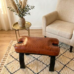 May include: A brown leather ottoman with a carved floral design sits on black legs. The ottoman is placed on a cream-colored rug with a black diamond pattern. A gold side table and beige armchair are in the background.