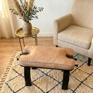 May include: A light brown leather ottoman with a geometric design on the top, set on black legs. The ottoman is placed on a cream-colored rug with a black diamond pattern. A vase of pampas grass sits on a small gold side table.