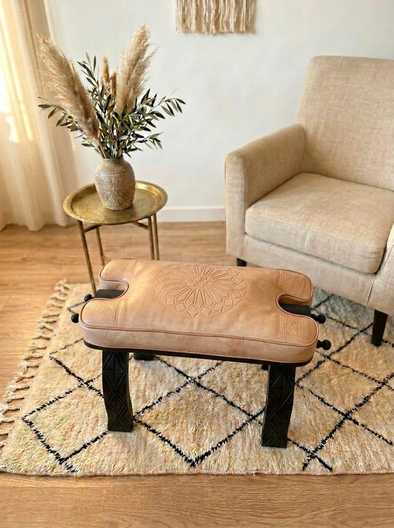 May include: A light brown leather ottoman with a geometric design on the top, set on black legs. The ottoman is placed on a cream-colored rug with a black diamond pattern. A vase of pampas grass sits on a small gold side table.