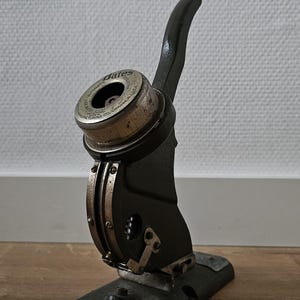 May include: An antique black and silver desk seal with a curved handle and a circular die. The die is marked "Bates Automatic" and "Orange, NJ, USA". The seal is mounted on a rectangular base and appears to be made of metal.