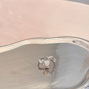 May include: A silver ring with a white opal stone set in a delicate, intricate design. The ring is on a silver tray.
