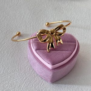 Puede incluir: Un brazalete dorado con lazo. El brazalete presenta un diseño de lazo detallado en el centro y pequeños detalles de bola en cada extremo. Se muestra sobre un joyero de terciopelo lavanda en forma de corazón.