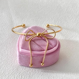 Puede incluir: Un brazalete dorado con diseño de lazo. El brazalete está sobre un joyero rosa en forma de corazón. El brazalete tiene una apariencia delicada y elegante, con un diseño simple pero elegante.