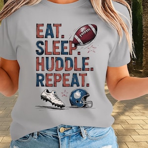 Puede incluir: Camiseta gris claro con las palabras "EAT. SLEEP. HUDDLE. REPEAT." en letras de bloque rojas y azules. La camiseta presenta un balón de fútbol, una bota de fútbol, una estrella y un casco de fútbol azul.