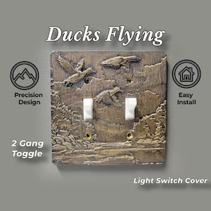 May include: A bronze-colored 2 gang toggle light switch cover featuring a raised relief design of ducks flying over a landscape with trees and water. The text "Ducks Flying" is printed above, with "Precision Design" and "Easy Install" labels.