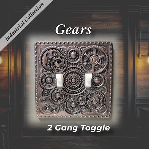 May include: A square 2 gang toggle light switch plate featuring a raised, metallic bronze-colored gear pattern. The plate includes the text Industrial Collection, Gears, and 2 Gang Toggle. Two white toggle switches are centered on the plate.
