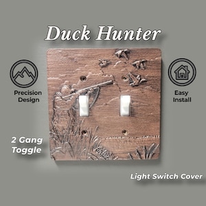 May include: A brown 2-gang toggle light switch cover featuring a raised relief design of a duck hunter aiming a shotgun at flying ducks. The text Duck Hunter, Precision Design, Easy Install, and 2 Gang Toggle Light Switch Cover appear.