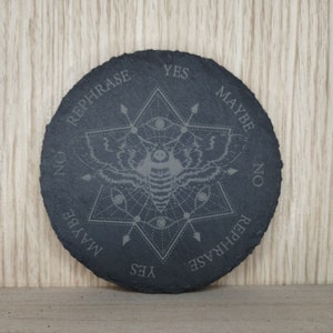 May include: Black slate round coaster with a geometric design featuring a moth and the words "Yes", "Maybe", "No", and "Rephrase" around the edge.
