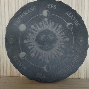 May include: A black slate circular divination board with a sun and moon design. The board has the words "Yes", "Maybe", "No", and "Rephrase" written around the edge.