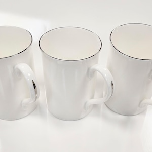 Puede incluir: Tres tazas de cerámica blanca con un borde plateado alrededor del borde superior y las asas. Las tazas están todas vacías y están sobre una superficie blanca.