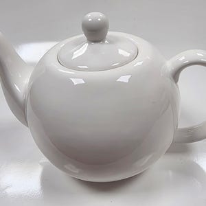 Puede incluir: Una tetera de cerámica blanca con cuerpo redondeado, boquilla curva y asa de bucle. La tapa tiene un pequeño pomo redondo. La tetera es brillante y refleja la luz.