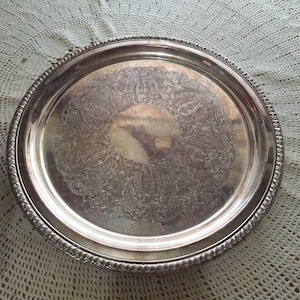 May include: A silver serving tray with a decorative embossed design. The tray has a raised rim and a smooth, reflective surface.