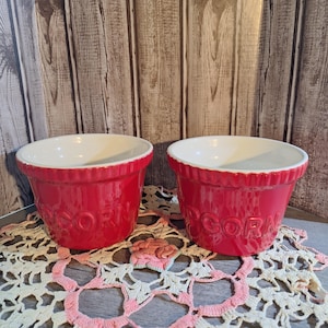 May include: Two red ceramic popcorn bowls with white interiors. The bowls have the word "Popcorn" embossed on the side.