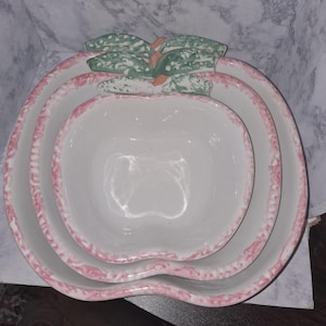 May include: Set of three ceramic apple-shaped bowls. The bowls are white with a pink speckled trim. The top of the apple has green leaves and a small orange stem. These decorative bowls are perfect for serving fruit or snacks.