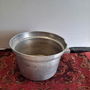 May include: A silver aluminum saucepan with a black handle. The saucepan has a rounded bottom and a wide, shallow rim.