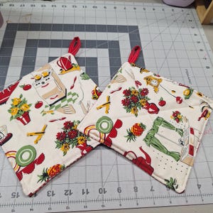 May include: Two quilted pot holders with a vintage kitchen-themed pattern. The fabric features red, yellow, and green kitchen utensils, appliances, and floral arrangements on a cream background. Each pot holder has a red loop for hanging.