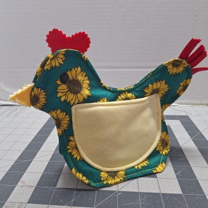May include: A handmade chicken-shaped pouch with a teal background and yellow sunflower pattern. It has a yellow pocket, a red felt comb, and red tail feathers. The beak is yellow.