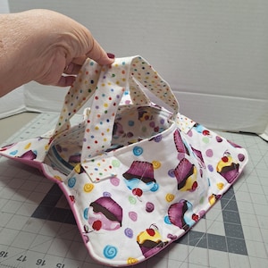 May include: A white fabric tote bag with a cupcake pattern. The bag features a pink trim and handles with colorful polka dots. The cupcake design includes purple frosting, cherries, and yellow frosting.
