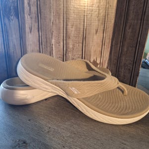 May include: A pair of beige Skechers flip-flops with a white sole. The flip-flops have a textured fabric strap and a contoured footbed. The Skechers logo is visible on the strap. The shoes are designed for comfort and casual wear.