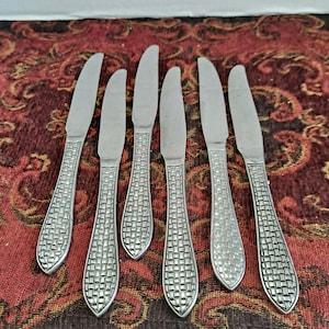 May include: Set of six silver dinner knives with a woven pattern on the handles. The knives are arranged in a row on a red and gold patterned surface.