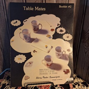 May include: A booklet titled "Table Mates" featuring a design with mugs, placemats, and a napkin. The cover includes the text "Booklet #2" and the name "Sherry Parker Summerfield." The booklet also displays the S&M Yarn Shop address and a price tag.