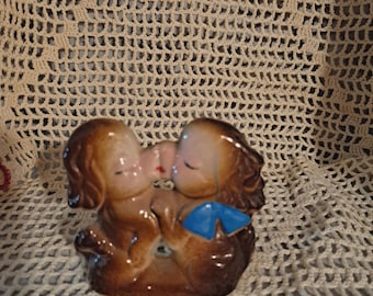 Könnte beinhalten: Vintage Keramikfigur, die zwei sich küssende Welpen mit braunem Haar darstellt. Ein Welpe hat eine blaue Schleife. Die Figur ist in Braun-, Beige- und Rosatönen bemalt. Die Welpen sind in einer liebevollen Umarmung.