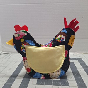 May include: A fabric chicken-shaped container with a black background and a pattern of cupcakes, strawberries, and flowers. It has a yellow pocket, a red comb, and a yellow beak. The container is designed to hold items.