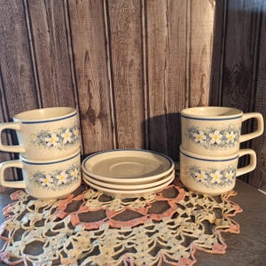 May include: Set of four floral teacups and saucers. The cups and saucers are white with a blue and yellow floral pattern. The cups are stacked on top of each other.
