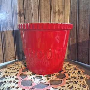 May include: A bright red popcorn bowl with the word "POP CORN" embossed on the side. The bowl has a ridged rim and is sitting on a decorative doily.