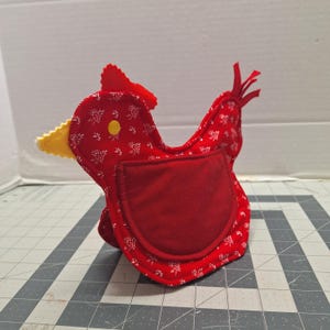 May include: A handmade red fabric chicken with white floral patterns. The chicken has a red comb, a yellow beak, and a red pocket. A yellow button is on the chicken's head. The chicken is sitting on a gray and white checkered surface.