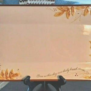 May include: Rectangular serving platter with a light beige surface and a brown rim. The corners are decorated with raised, golden-yellow leaf designs. The phrase "Give us this day our daily bread" is written in script along the bottom edge.